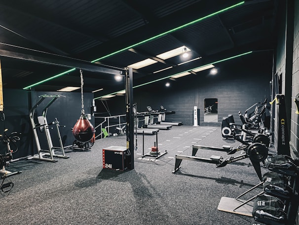 A modern gym interior equipped with various fitness machines such as treadmills, rowing machines, and a punching bag. The room has a dark color scheme with green accent lighting running across the ceiling and walls. The floor is carpeted, and there's a sense of tidy organization.