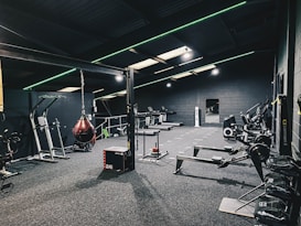 A modern gym interior equipped with various fitness machines such as treadmills, rowing machines, and a punching bag. The room has a dark color scheme with green accent lighting running across the ceiling and walls. The floor is carpeted, and there's a sense of tidy organization.