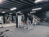 The image depicts a modern gym interior with various exercise equipment including weight racks, benches, and machines. The gym is well-lit with fluorescent lights, and the walls are adorned with mirrors. There are dumbbells neatly stacked in racks, and a staircase is visible leading to an overhead area.