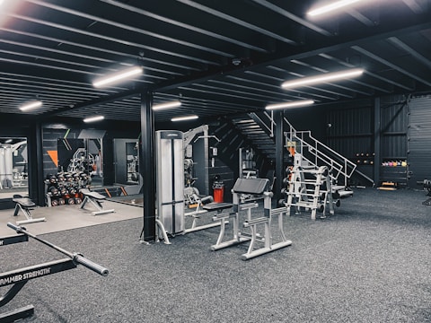 The image depicts a modern gym interior with various exercise equipment including weight racks, benches, and machines. The gym is well-lit with fluorescent lights, and the walls are adorned with mirrors. There are dumbbells neatly stacked in racks, and a staircase is visible leading to an overhead area.