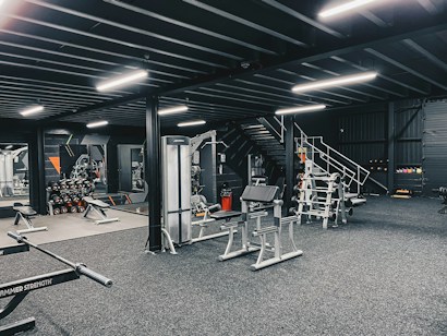 The image depicts a modern gym interior with various exercise equipment including weight racks, benches, and machines. The gym is well-lit with fluorescent lights, and the walls are adorned with mirrors. There are dumbbells neatly stacked in racks, and a staircase is visible leading to an overhead area.