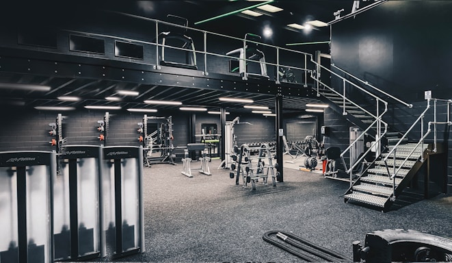 A modern gym interior with various exercise equipment, including weight racks, treadmills, and other fitness machines. The space is well-lit with industrial-style lighting, and features a mezzanine level accessed by metal stairs.