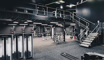 A modern gym interior with various exercise equipment, including weight racks, treadmills, and other fitness machines. The space is well-lit with industrial-style lighting, and features a mezzanine level accessed by metal stairs.