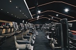 A spacious gym filled with various exercise equipment. Treadmills are aligned in rows on one side, while weight machines and other fitness devices are organized on the other side. The ceiling features modern, angular lines with bright overhead lights, adding a sleek and contemporary feel.