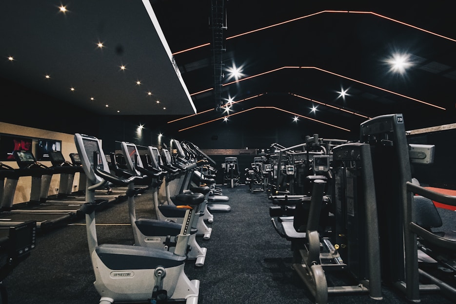 A spacious gym filled with various exercise equipment. Treadmills are aligned in rows on one side, while weight machines and other fitness devices are organized on the other side. The ceiling features modern, angular lines with bright overhead lights, adding a sleek and contemporary feel.