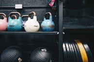 A collection of kettlebells in various colors, including pink, blue, white, and purple, are neatly arranged on a black metal shelf. The kettlebells are marked with their respective weights in kilograms. Below the shelf, two black medicine balls are visible, also marked with their weight. To the side, a series of stacked weight plates can be seen in black and yellow. The setting suggests a gym environment with an industrial appearance due to the dark wall and metal rack.