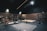 A dimly lit gym with wooden walls featuring weightlifting platforms and various weight racks. Plates with different weight denominations are neatly stacked on the floor and weightlifting bars are set in place for use. The ceiling lights illuminate specific areas, providing a focused ambiance for heavy lifting.