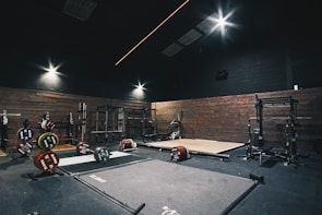 A dimly lit gym with wooden walls featuring weightlifting platforms and various weight racks. Plates with different weight denominations are neatly stacked on the floor and weightlifting bars are set in place for use. The ceiling lights illuminate specific areas, providing a focused ambiance for heavy lifting.