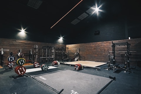 A dimly lit gym with wooden walls featuring weightlifting platforms and various weight racks. Plates with different weight denominations are neatly stacked on the floor and weightlifting bars are set in place for use. The ceiling lights illuminate specific areas, providing a focused ambiance for heavy lifting.