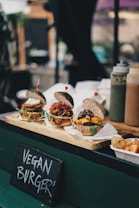Three gourmet vegan burgers are artfully arranged on a wooden serving board. Each burger is topped with a variety of colorful vegetables and sauces. The buns have a rustic look, with seeds visible on the surface. In the background, several condiment bottles are placed, suggesting a food stall or a street food setting. A small sign in front reads 'Vegan Burgers', indicating that these are plant-based options.