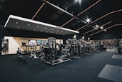A spacious gym interior featuring various exercise machines, including treadmills, elliptical trainers, and weight lifting equipment. The dark ceiling is accentuated with orange LED strip lighting, creating an ambient atmosphere. The gym is well-lit and clean, with a modern design and advertisement posters on the walls.