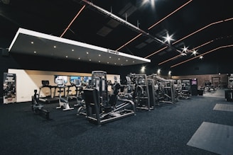 A spacious gym interior featuring various exercise machines, including treadmills, elliptical trainers, and weight lifting equipment. The dark ceiling is accentuated with orange LED strip lighting, creating an ambient atmosphere. The gym is well-lit and clean, with a modern design and advertisement posters on the walls.