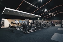 A spacious gym interior featuring various exercise machines, including treadmills, elliptical trainers, and weight lifting equipment. The dark ceiling is accentuated with orange LED strip lighting, creating an ambient atmosphere. The gym is well-lit and clean, with a modern design and advertisement posters on the walls.
