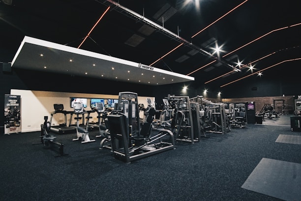 A spacious gym interior featuring various exercise machines, including treadmills, elliptical trainers, and weight lifting equipment. The dark ceiling is accentuated with orange LED strip lighting, creating an ambient atmosphere. The gym is well-lit and clean, with a modern design and advertisement posters on the walls.