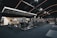 A spacious gym interior featuring various exercise machines, including treadmills, elliptical trainers, and weight lifting equipment. The dark ceiling is accentuated with orange LED strip lighting, creating an ambient atmosphere. The gym is well-lit and clean, with a modern design and advertisement posters on the walls.