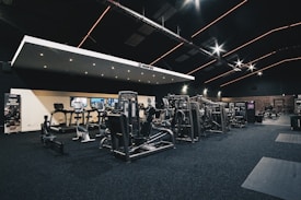 A spacious gym interior featuring various exercise machines, including treadmills, elliptical trainers, and weight lifting equipment. The dark ceiling is accentuated with orange LED strip lighting, creating an ambient atmosphere. The gym is well-lit and clean, with a modern design and advertisement posters on the walls.