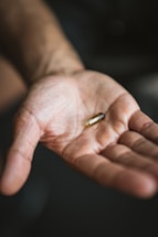 A close-up of an open hand with a single, transparent gel capsule resting on the palm. The background is softly blurred, emphasizing the focus on the hand and pill.