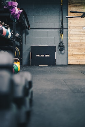 A gym setting featuring a variety of workout equipment. Resistance bands and TRX straps are hanging on the wall. Kettlebells in different colors are placed on a rack, and a box labeled 'Bulldog Gear' is situated against a gray brick wall. The floor is covered with black gym mats, and there's a wooden panel on the right side.