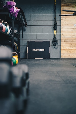 A gym setting featuring a variety of workout equipment. Resistance bands and TRX straps are hanging on the wall. Kettlebells in different colors are placed on a rack, and a box labeled 'Bulldog Gear' is situated against a gray brick wall. The floor is covered with black gym mats, and there's a wooden panel on the right side.