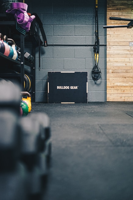 A gym setting featuring a variety of workout equipment. Resistance bands and TRX straps are hanging on the wall. Kettlebells in different colors are placed on a rack, and a box labeled 'Bulldog Gear' is situated against a gray brick wall. The floor is covered with black gym mats, and there's a wooden panel on the right side.
