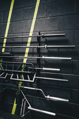 A set of gym equipment, specifically various bars and handles, organized on a black wall with green diagonal stripes painted across it. The equipment appears to be neatly arranged, showcasing different types of bars used for weightlifting.