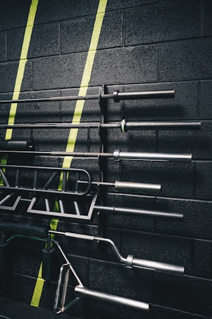 A set of gym equipment, specifically various bars and handles, organized on a black wall with green diagonal stripes painted across it. The equipment appears to be neatly arranged, showcasing different types of bars used for weightlifting.