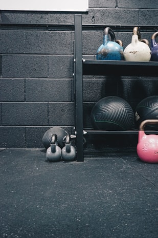Kettlebells of different colors and sizes are arranged on a black metal shelf against a textured black brick wall. Below the shelf, there are two smaller black kettlebells on the floor along with a large exercise ball. The floor appears to be covered in black gym flooring with a slightly rough texture.