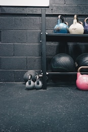 Kettlebells of different colors and sizes are arranged on a black metal shelf against a textured black brick wall. Below the shelf, there are two smaller black kettlebells on the floor along with a large exercise ball. The floor appears to be covered in black gym flooring with a slightly rough texture.