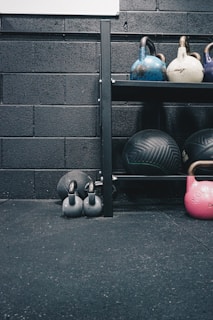 Kettlebells of different colors and sizes are arranged on a black metal shelf against a textured black brick wall. Below the shelf, there are two smaller black kettlebells on the floor along with a large exercise ball. The floor appears to be covered in black gym flooring with a slightly rough texture.