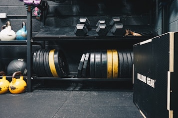A gym setting featuring various fitness equipment, including kettlebells, stacked weight plates on a rack, dumbbells, and a medicine ball. The gym has a rugged, industrial vibe with black walls and a Bulldog Gear plyo box.