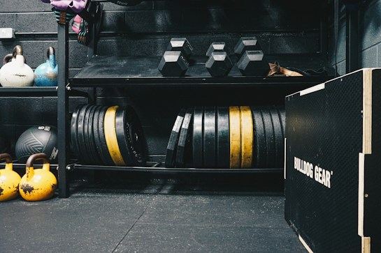 A gym setting featuring various fitness equipment, including kettlebells, stacked weight plates on a rack, dumbbells, and a medicine ball. The gym has a rugged, industrial vibe with black walls and a Bulldog Gear plyo box.