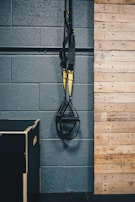 A pair of black and yellow suspension training straps hang from a sturdy metal support against a dark gray textured wall. A wooden panelled section is adjacent, with a black exercise box positioned on the floor.