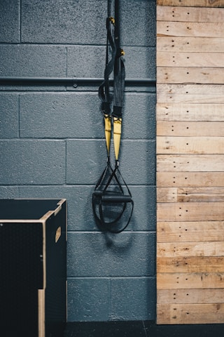 A pair of black and yellow suspension training straps hang from a sturdy metal support against a dark gray textured wall. A wooden panelled section is adjacent, with a black exercise box positioned on the floor.