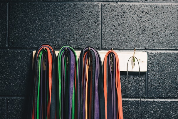 Multiple resistance bands of various colors, including red, green, purple, and orange, are neatly hung on a white wall-mounted rack against a textured, dark wall. The bands are looped over metal hooks.