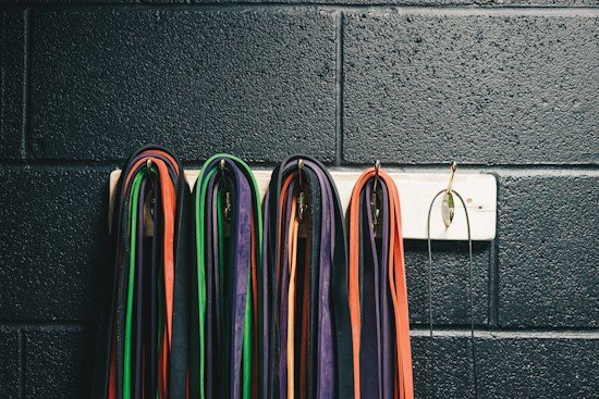 Multiple resistance bands of various colors, including red, green, purple, and orange, are neatly hung on a white wall-mounted rack against a textured, dark wall. The bands are looped over metal hooks.