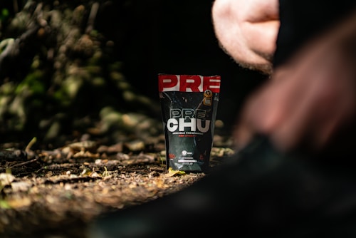 A package of a dietary supplement is prominently positioned on the forest floor, surrounded by dry leaves and blurred natural elements in the background. Partial views of a person appear in the foreground, suggesting outdoor activity.