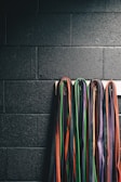 A collection of martial arts belts in various colors, including black, orange, green, purple, and red, hanging down a dark brick wall.