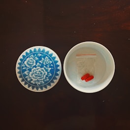 A decorative ceramic dish with a floral blue and white pattern lies next to an open bowl containing two red capsules in a small transparent ziplock bag, placed on a dark wooden surface.