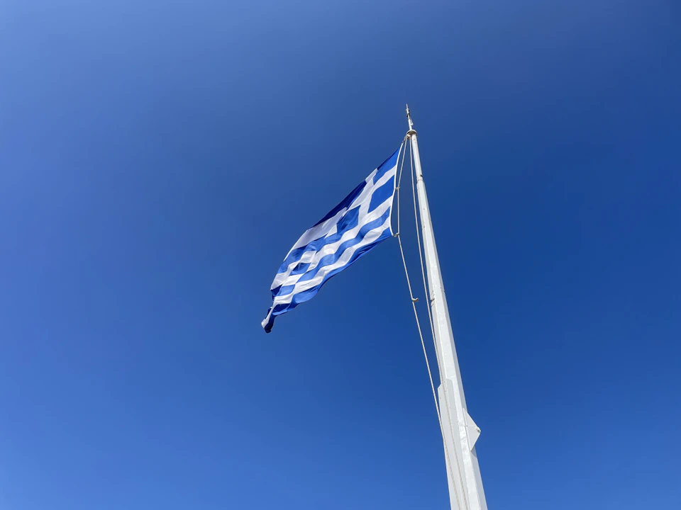 The image shows a Greek flag flying high against a clear blue sky. The flag is attached to a white pole and displays the traditional Greek design with blue, white, and black stripes on its horizontal bands.