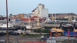 A harbor scene with multiple fishing boats docked near an industrial area. The background features large warehouse buildings, various machinery, and cranes. The foreground includes a construction vehicle labeled 'REUSE' and some scattered equipment and materials.