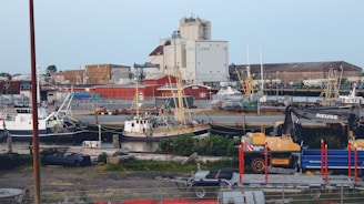 A harbor scene with multiple fishing boats docked near an industrial area. The background features large warehouse buildings, various machinery, and cranes. The foreground includes a construction vehicle labeled 'REUSE' and some scattered equipment and materials.