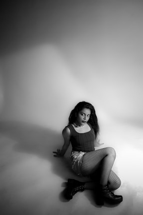 A woman is seated on the floor in a studio setting, wearing a sleeveless top, fishnet stockings, and boots. The lighting is soft, creating shadows on the backdrop and floor, highlighting the monochromatic theme of the photograph.