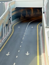 A smooth, empty road leads into a tunnel with two bright yellow lines and painted murals on the walls. Arrows are marked on the asphalt to guide traffic direction, and the curve of the tunnel creates a sense of anticipation.
