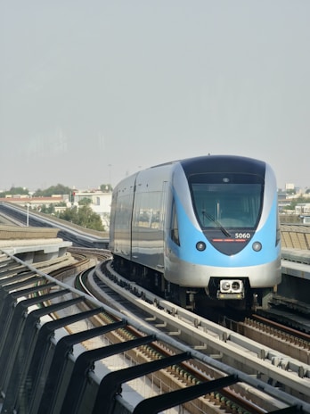 A modern train with a sleek design travels along an elevated track. Urban buildings and greenery are visible in the distance under a clear sky.