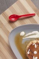 A partially visible dessert, likely a flan, sits on a round gray plate. The flan is garnished with a caramel sauce and small dollops of cream, with some sauce spreading onto the plate. A vibrant red spoon is placed on a wooden surface beside the plate, enhancing the visual contrast.