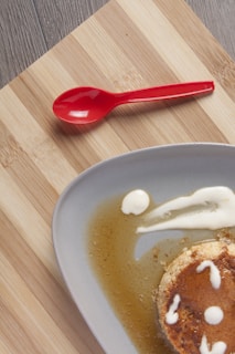 A partially visible dessert, likely a flan, sits on a round gray plate. The flan is garnished with a caramel sauce and small dollops of cream, with some sauce spreading onto the plate. A vibrant red spoon is placed on a wooden surface beside the plate, enhancing the visual contrast.