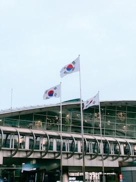Three South Korean flags are prominently displayed, fluttering in front of a modern building with a curved, glass facade. The building appears to be large and might serve a public or governmental function. The sky is overcast with a light blue hue.