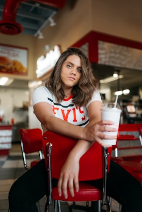 A person with medium-length brown hair is seated in a retro-style diner, resting their arms on the back of a red chair and holding a white milkshake. The diner features red and white decor, with checkerboard floor tiles. There is a well-lit area in the background with a sign and a counter.