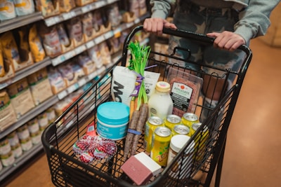 A shopping cart filled with various grocery items such as vegetables, canned drinks, a carton of milk, deli meat, and packaged snacks. The cart is being pushed by a person wearing a gray top and patterned pants in a grocery store aisle.