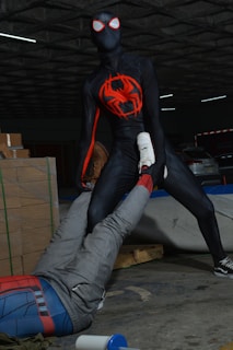 A person wearing a black spider-themed superhero costume with red accents stands in an assertive pose, holding the leg of another person dressed in a Spider-Man costume. The scene takes place in what appears to be a dimly lit warehouse with stacked cardboard boxes and a few visible parked cars.